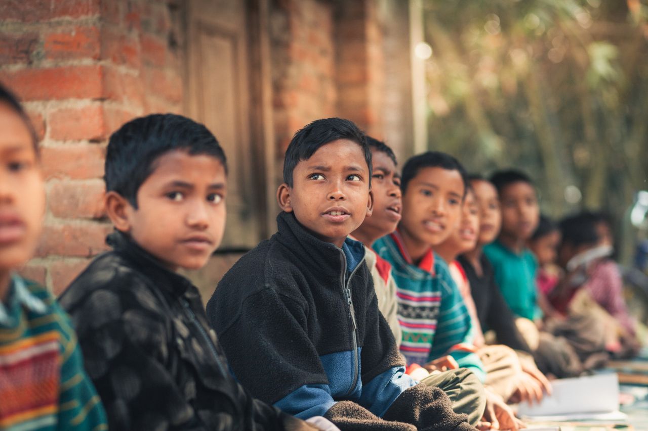 gallery-1 A group of children from Bangladesh sitting outdoors, engaged in learning at a rural school.